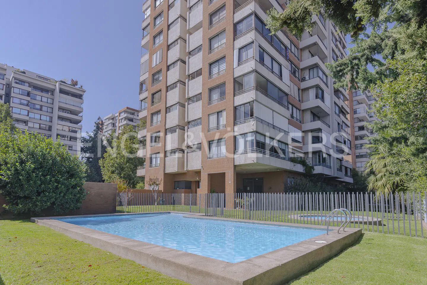 Outdoor pool with blue water, surrounded by green grass and a white fence, in front of a tall apartment building.