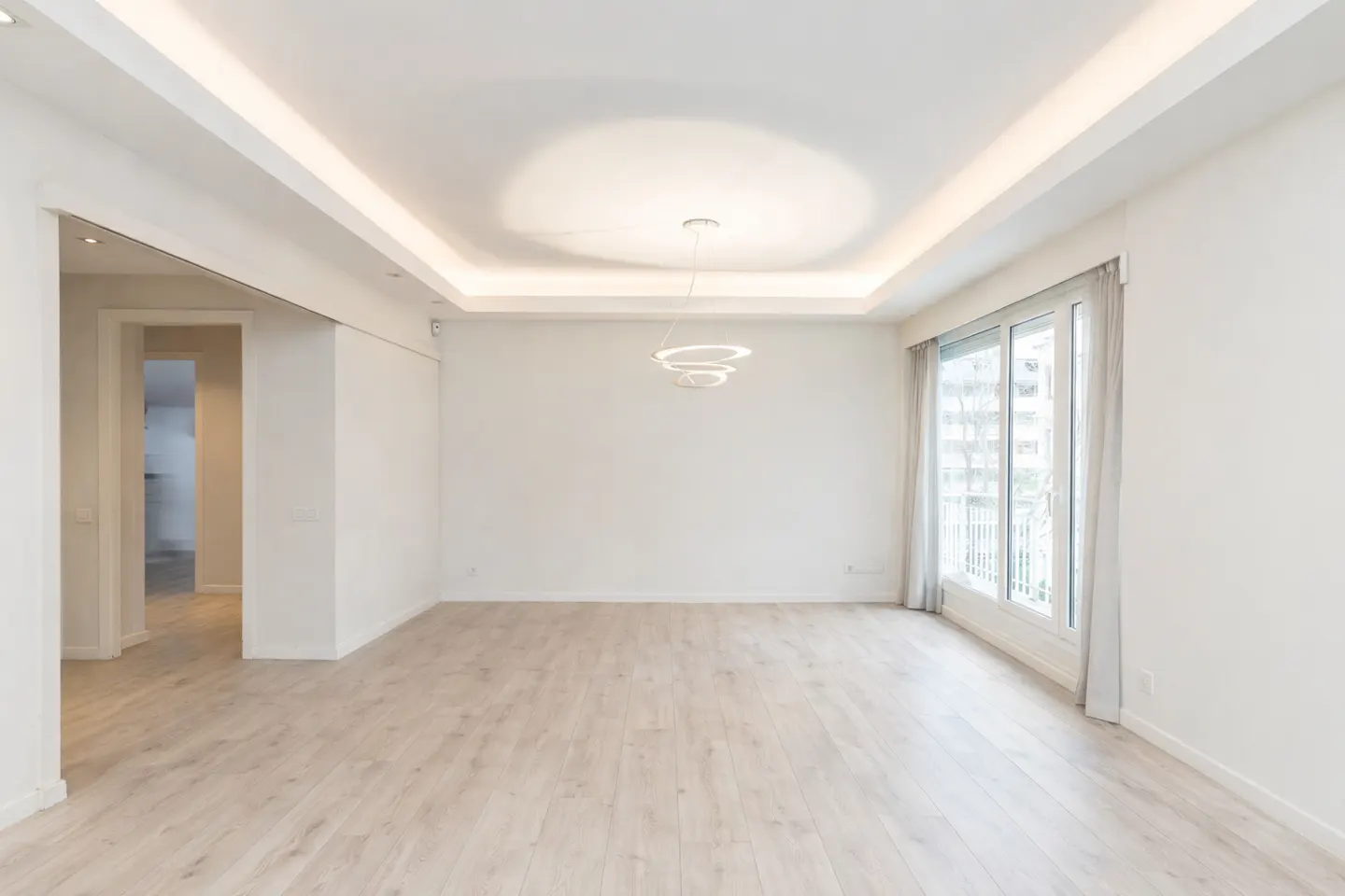Bright, empty room with light wood floors, white walls, and a modern chandelier. Large windows offer a city view.