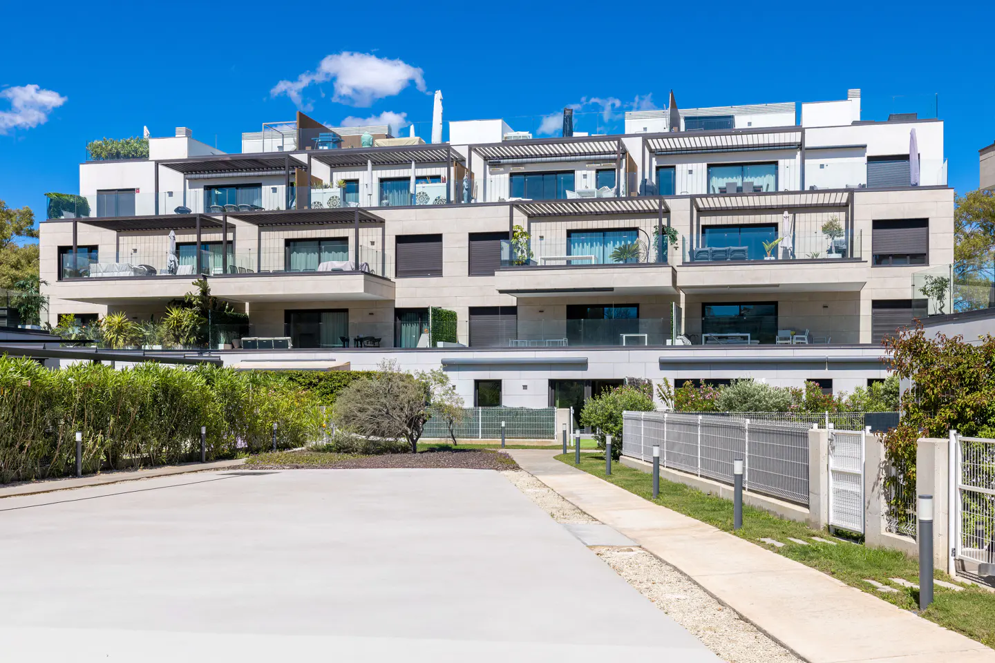 Modern multi-story townhouses with balconies, stone facades, and rooftop terraces under a blue sky. Green hedges and trees surround the property.