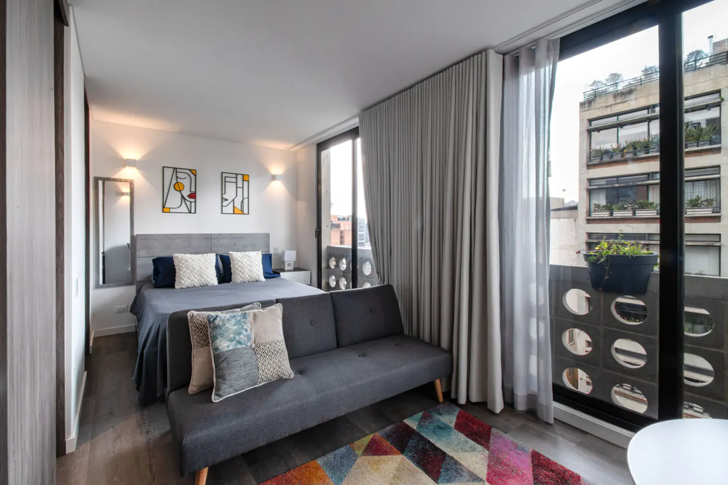 Bedroom with a gray sofa, bed, and balcony. Abstract art hangs above the bed. A colorful rug is on the floor.