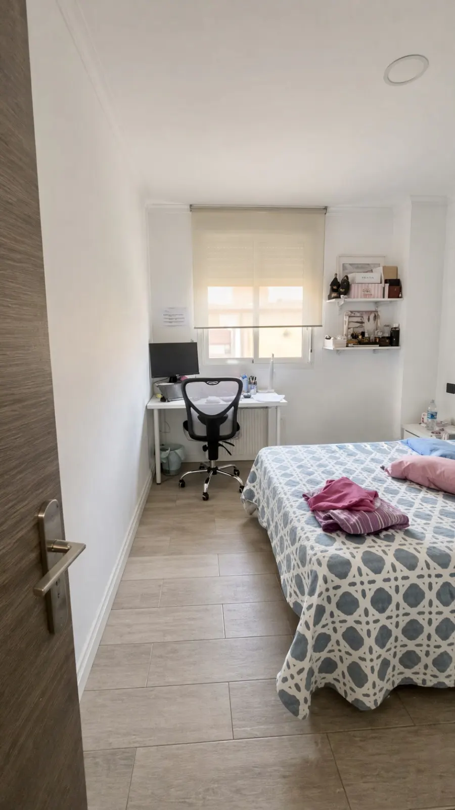 A bedroom with a desk, chair, and bed. The bed has a blue and white patterned cover. A computer sits on the desk by the window.