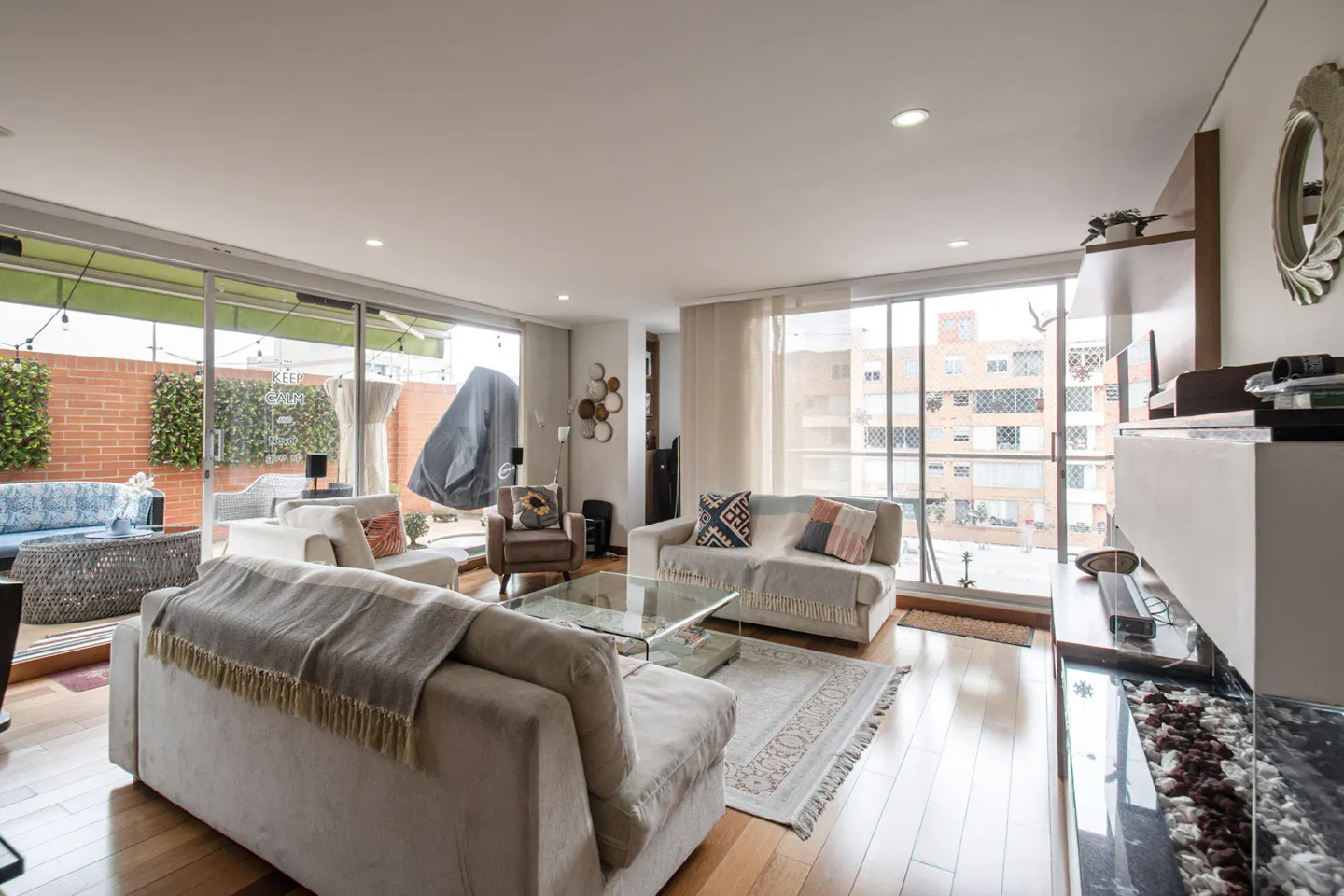 Bright living room with hardwood floors, sofas, glass table, and large windows leading to a patio with greenery.