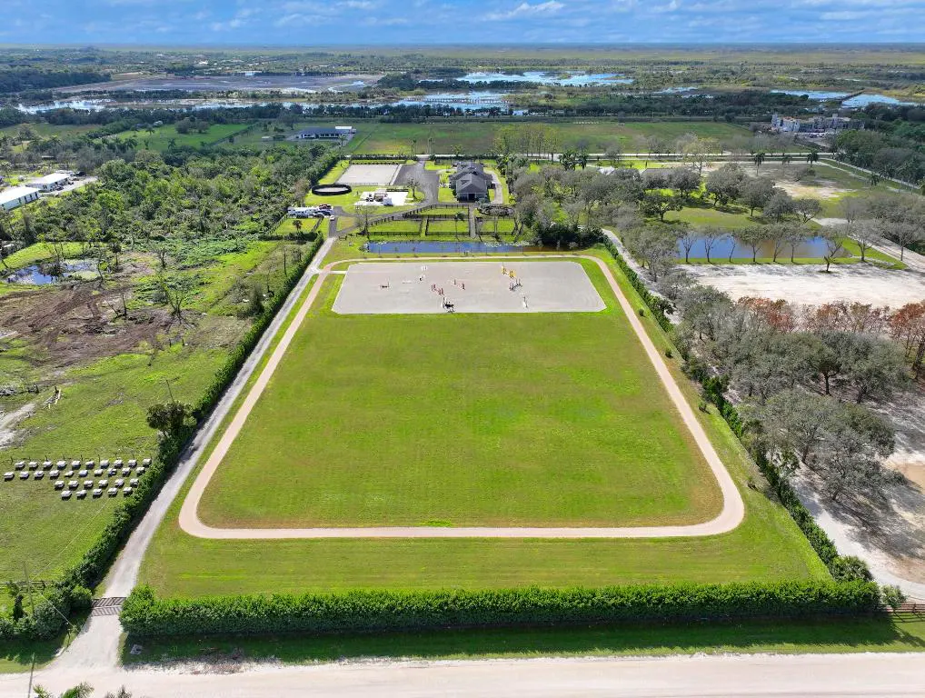 Aerial view of a horse training facility with a large green field, sand arena, stables, and surrounding trees.