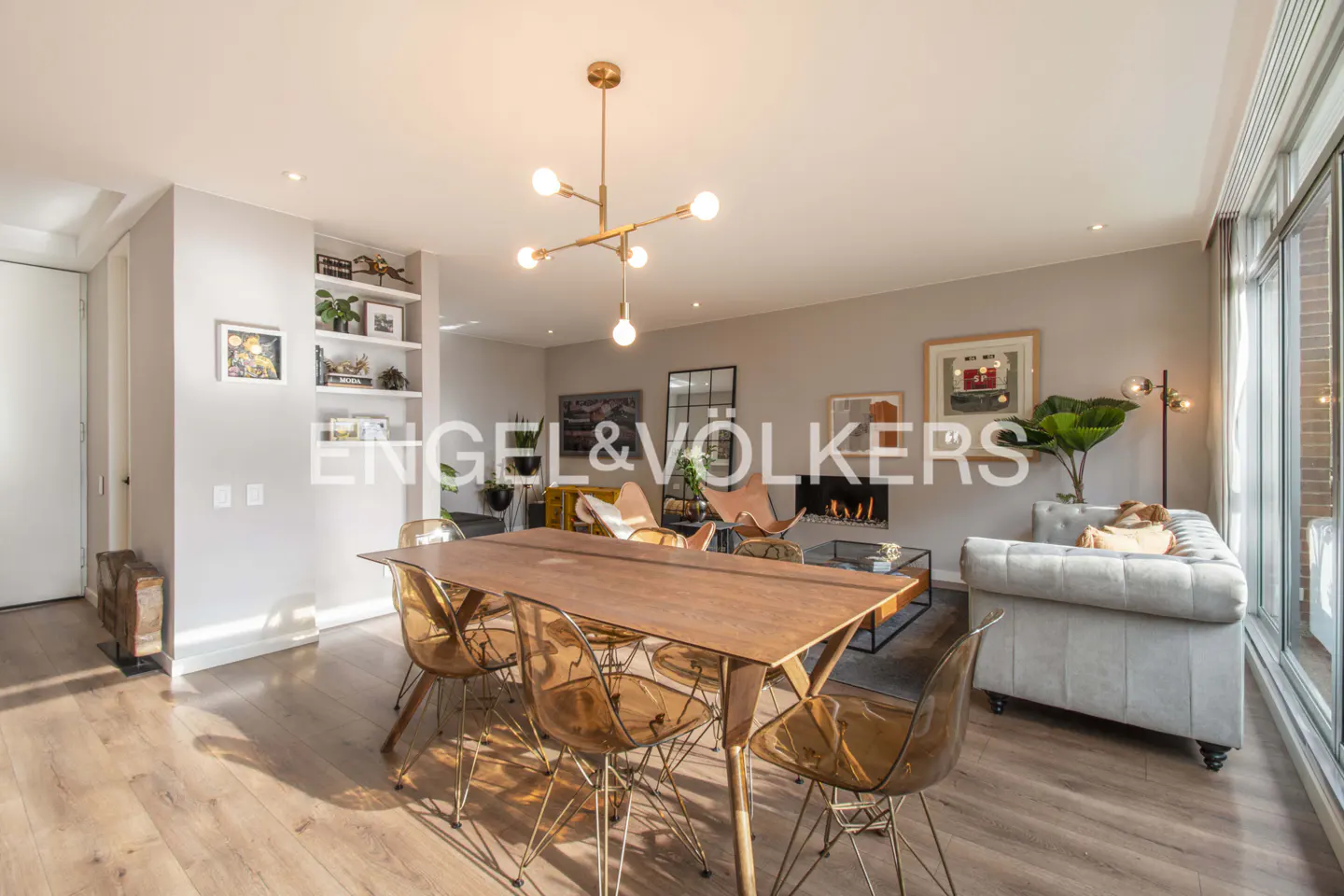 Open-concept living and dining area with wood floors, a fireplace, and a gray sofa. A wooden table with clear chairs sits under a modern chandelier.