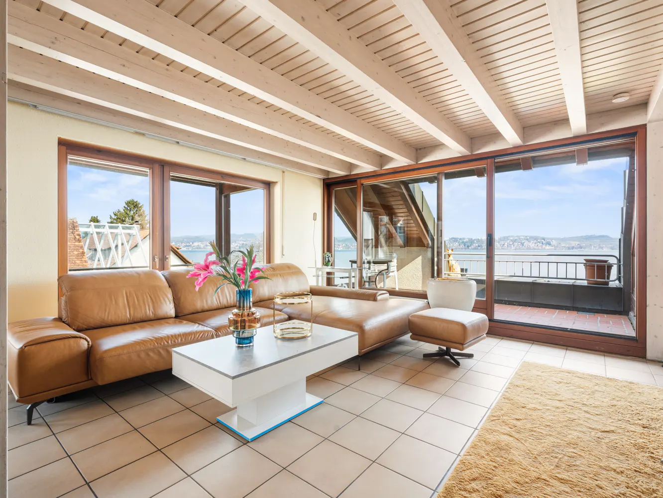 Living room with brown leather sofa, white coffee table, and large windows overlooking a lake. The ceiling has exposed wooden beams.