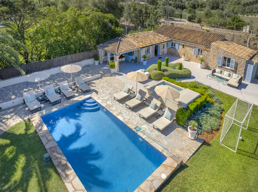 Aerial view of a luxury villa with a blue pool, lounge chairs, umbrellas, and a soccer goal on a green lawn. The villa has a stone facade and a tiled roof.