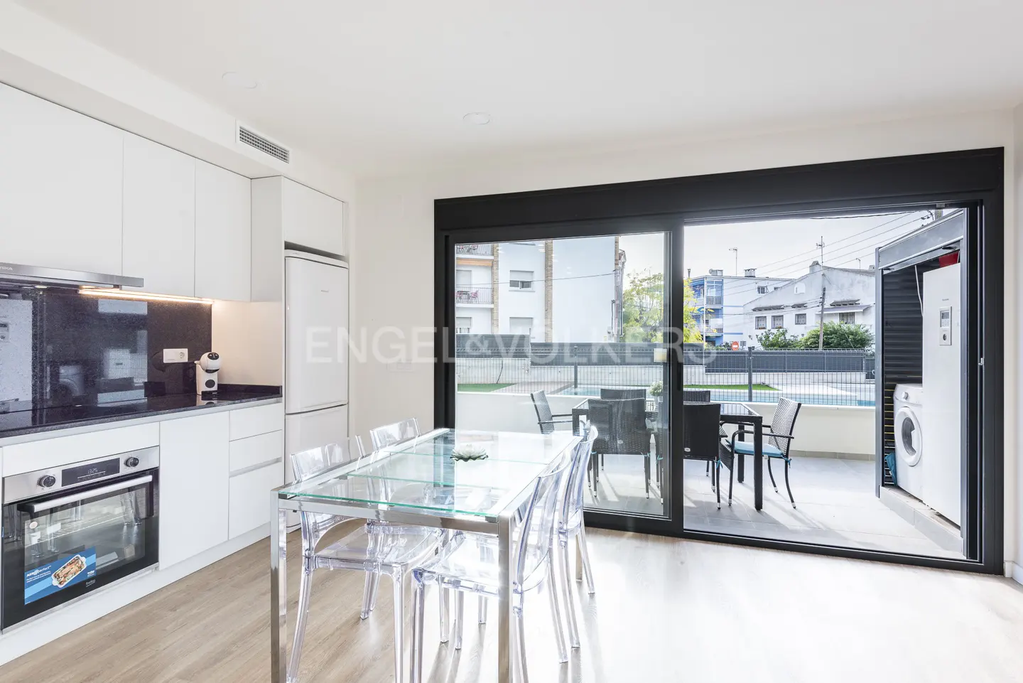Bright kitchen with white cabinets, black countertops, and a glass table with clear chairs. Sliding doors open to a patio with outdoor seating and a pool.