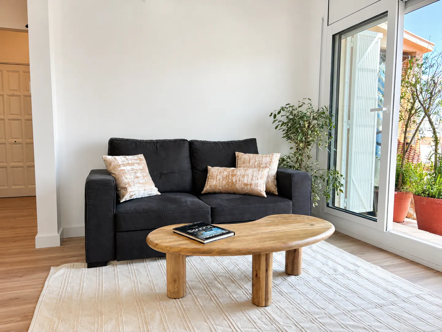 Bright living room with a black sofa, patterned pillows, and a wooden coffee table on a striped rug, next to a balcony.