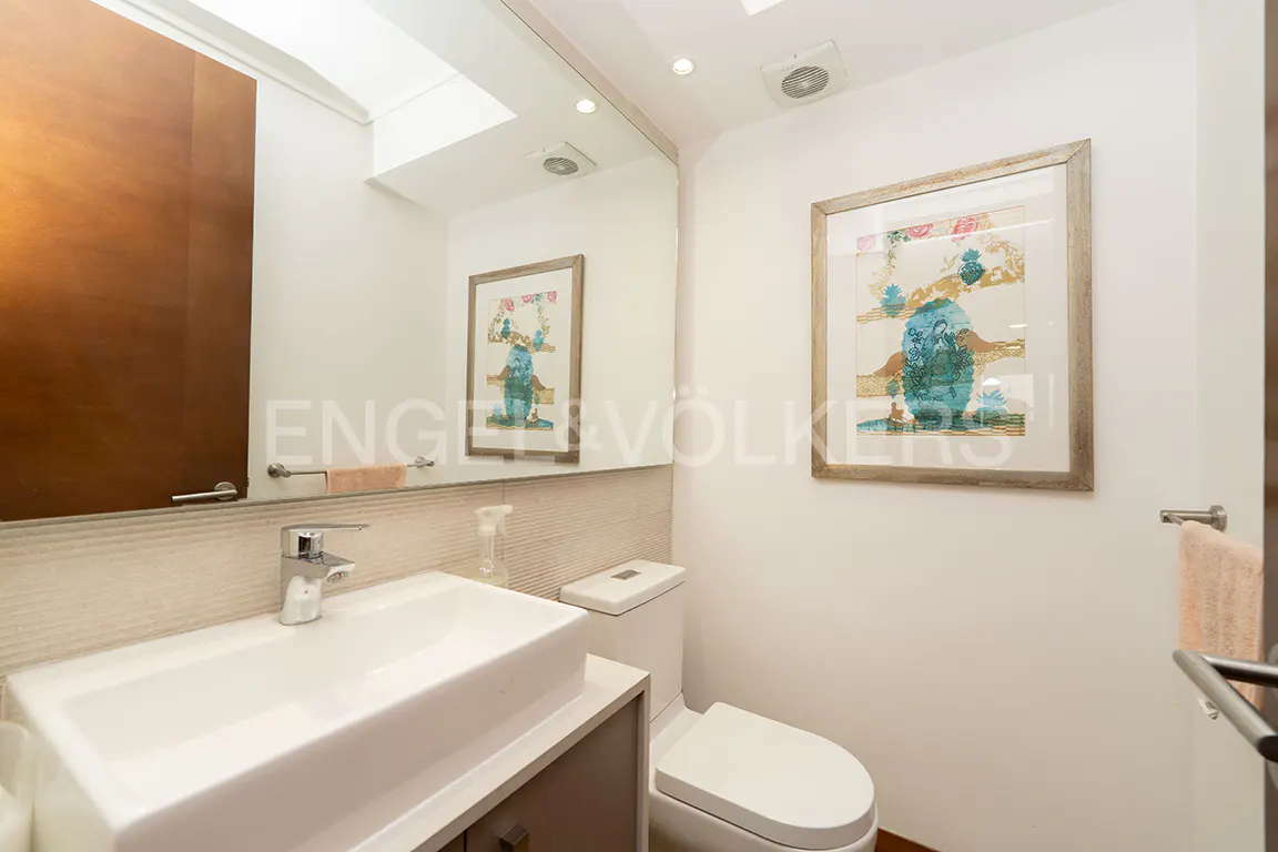 Modern bathroom with white sink, toilet, and brown cabinet. Artwork hangs on the white wall. A large mirror reflects the scene.