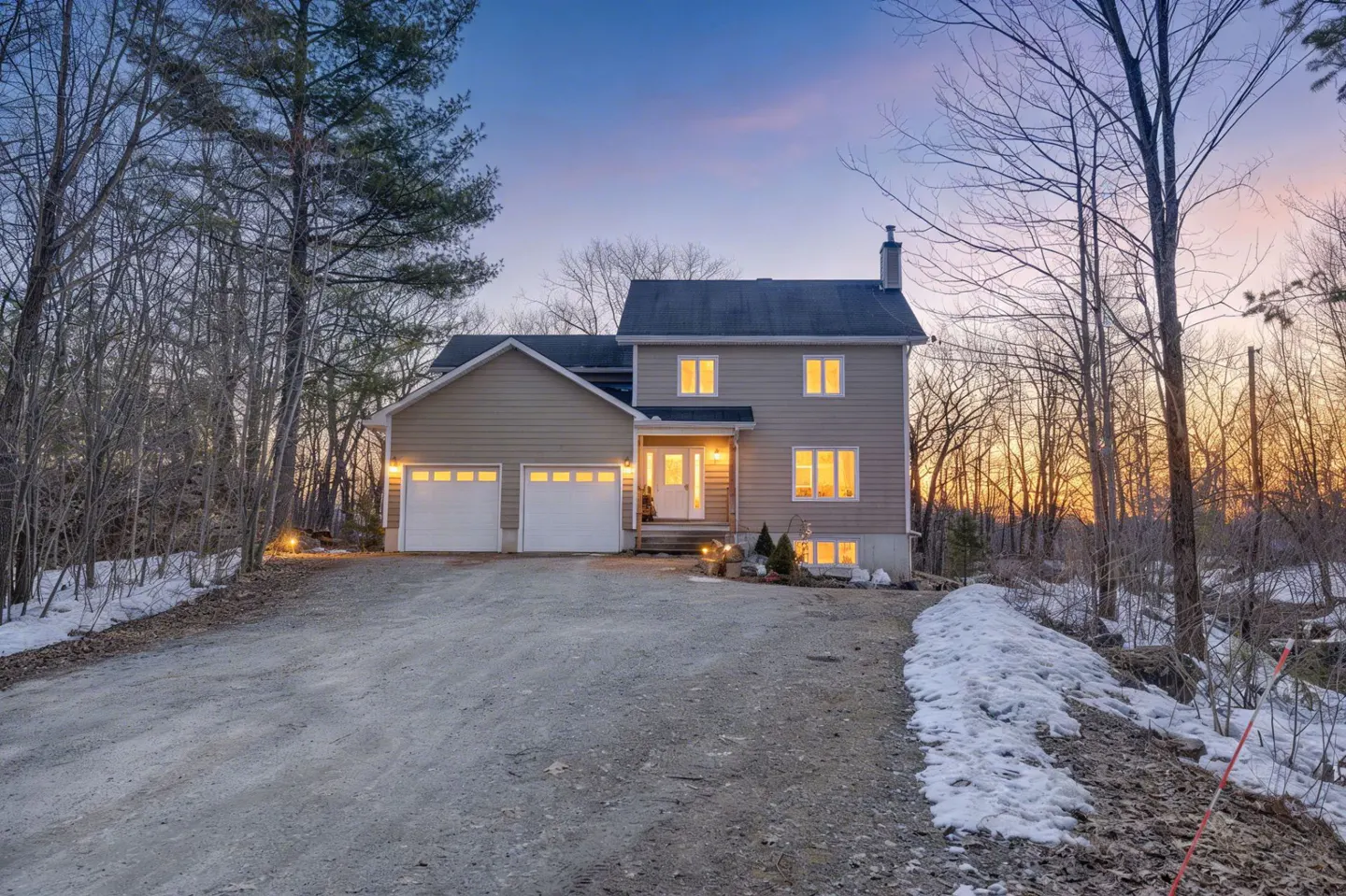 Two-story beige house with a two-car garage at dusk, surrounded by bare trees and patches of snow on the ground.