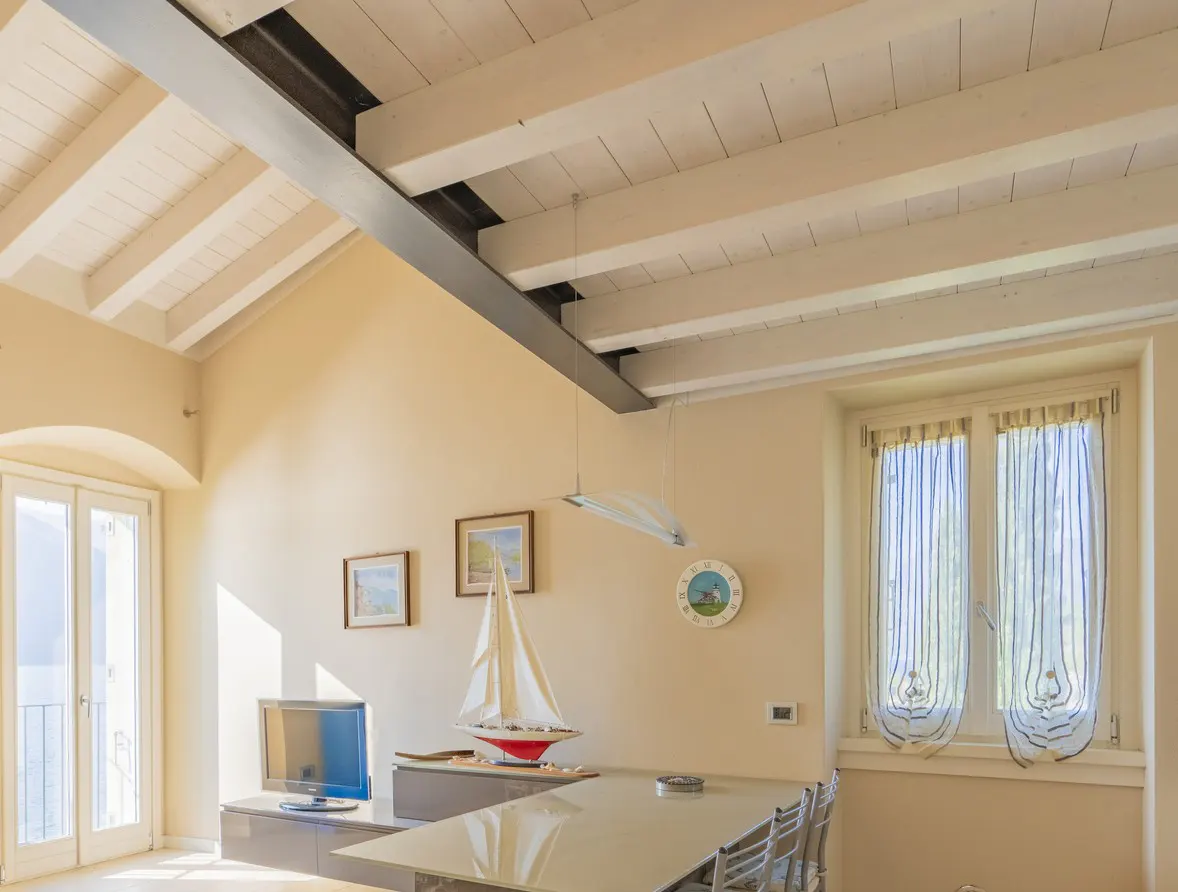 Bright living room with white beamed ceiling, beige walls, and a window with blue striped curtains. A sailboat model sits on a cabinet.