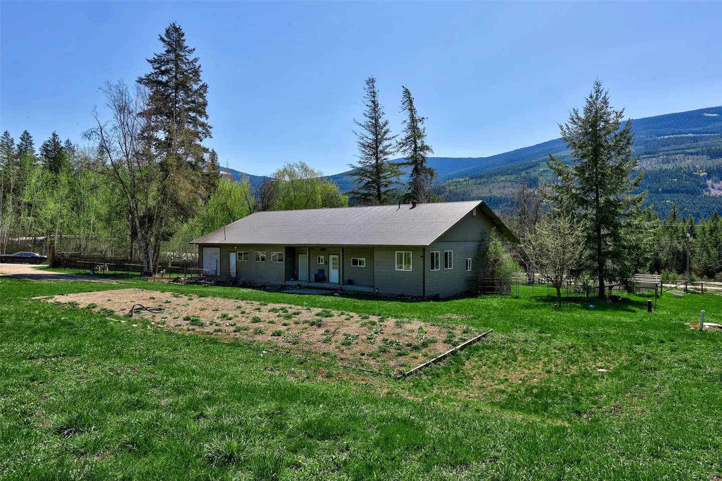 A single-story, gray house with a metal roof sits on a green lawn with trees and mountains in the background.