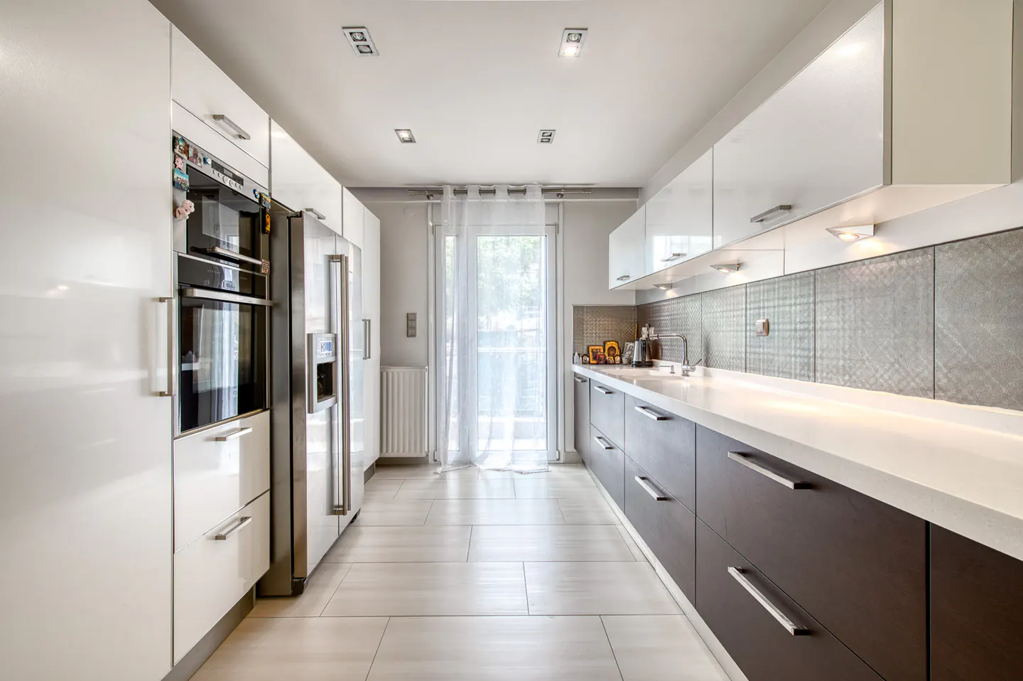A modern kitchen with white cabinets, stainless steel appliances, and dark brown lower cabinets. A window with sheer curtains is at the end of the room.