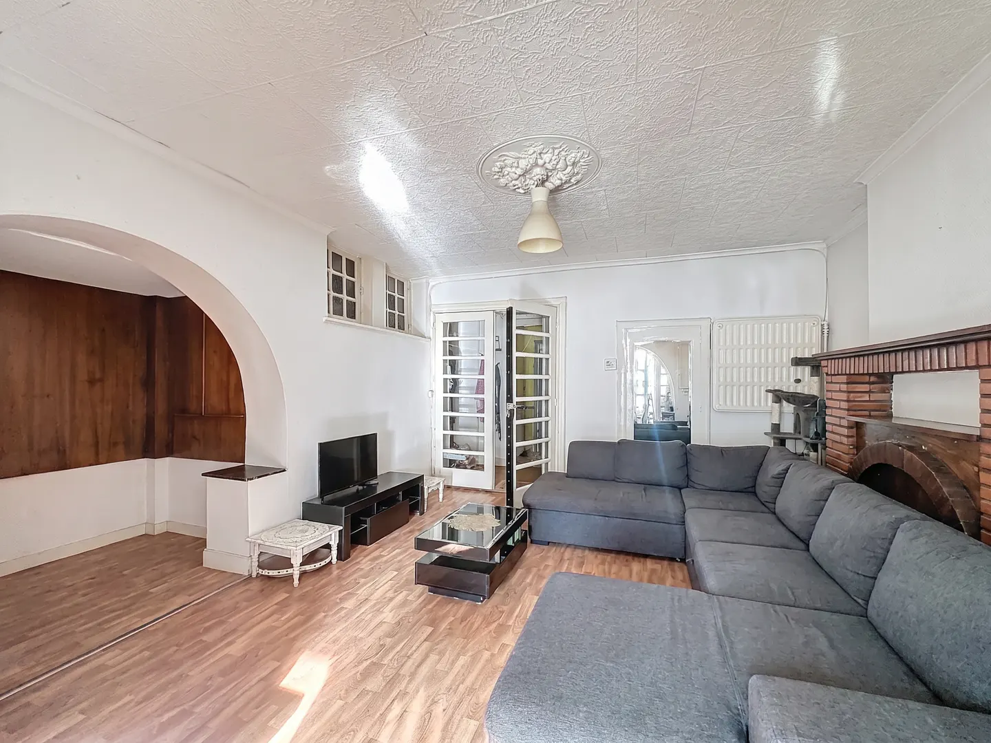 Bright living room with wood floors, white walls, and a gray sectional sofa. A TV sits on a black stand near an arched doorway. French doors lead to another room.
