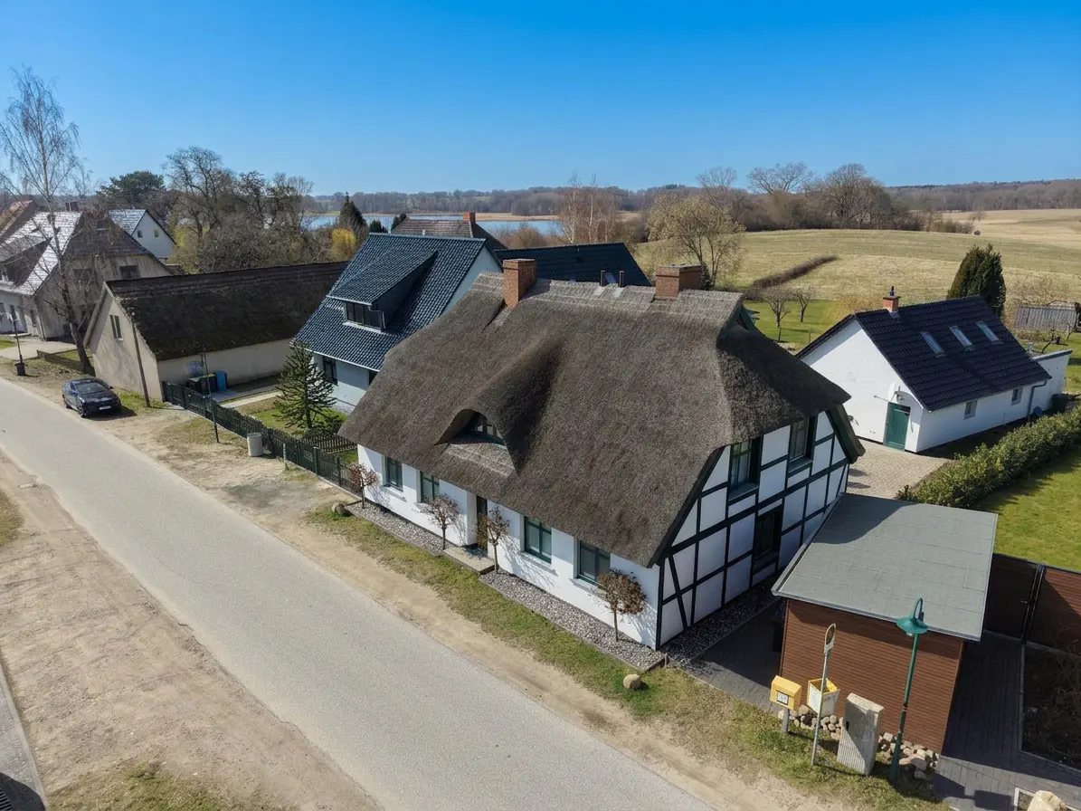 Aerial view of a white, half-timbered house with a thatched roof on a sunny day. Other houses and a road are visible.