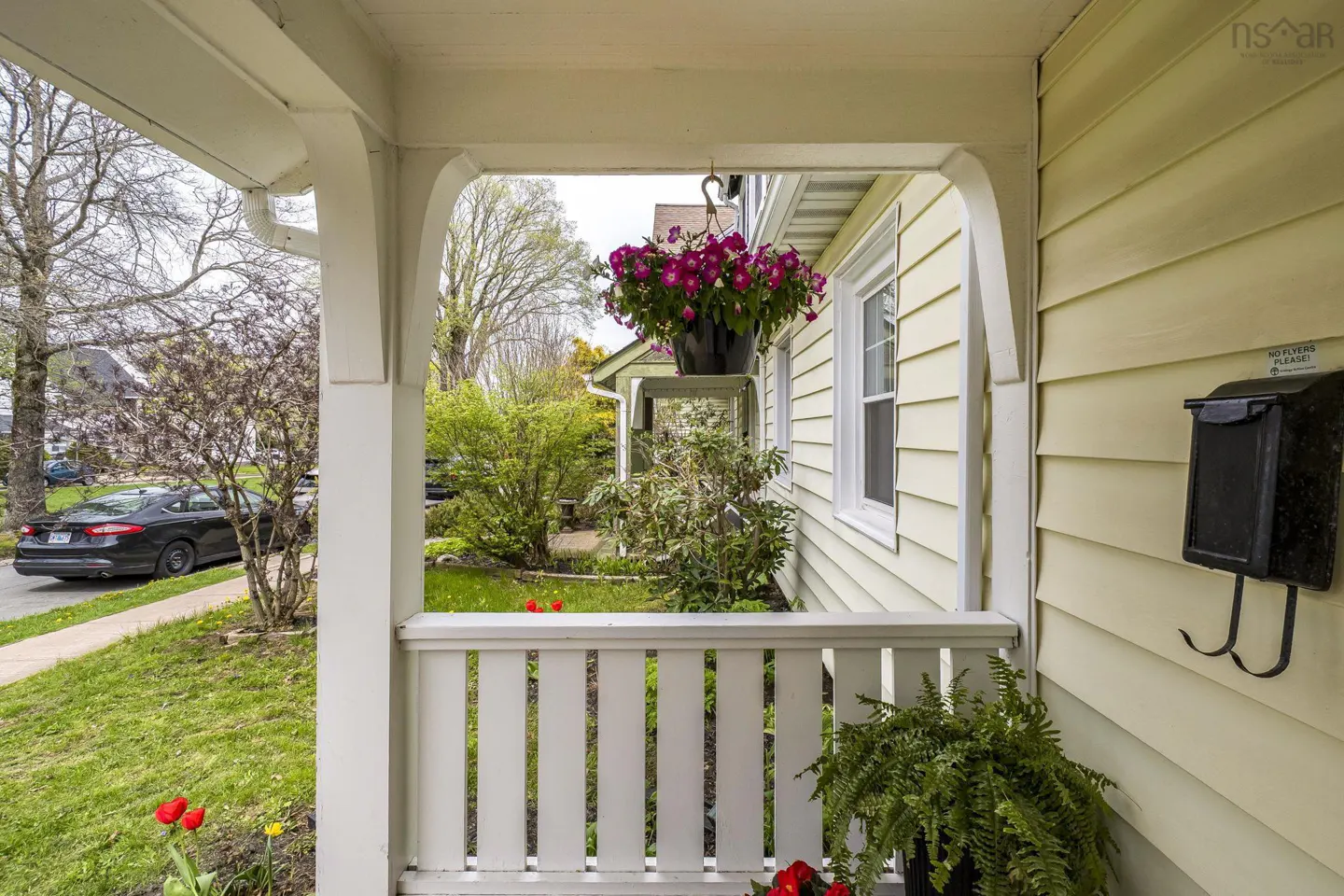 View from a porch with white railings, a hanging flower basket, and a black mailbox on a yellow house.