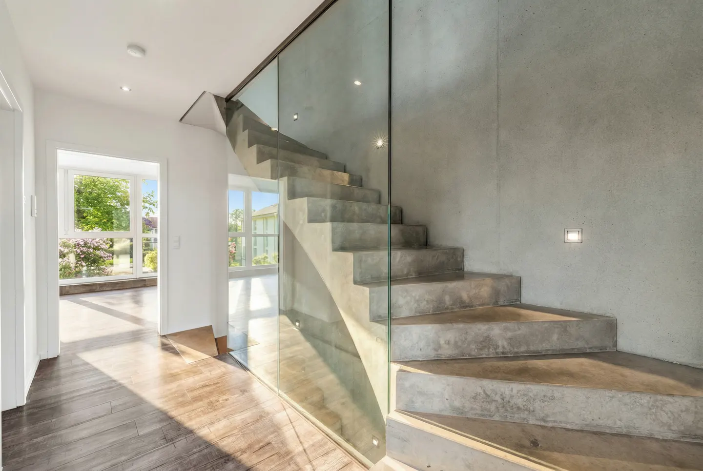 Modern home interior with concrete stairs, glass railing, and wood floors. Sunlight streams through a doorway to a bright room.