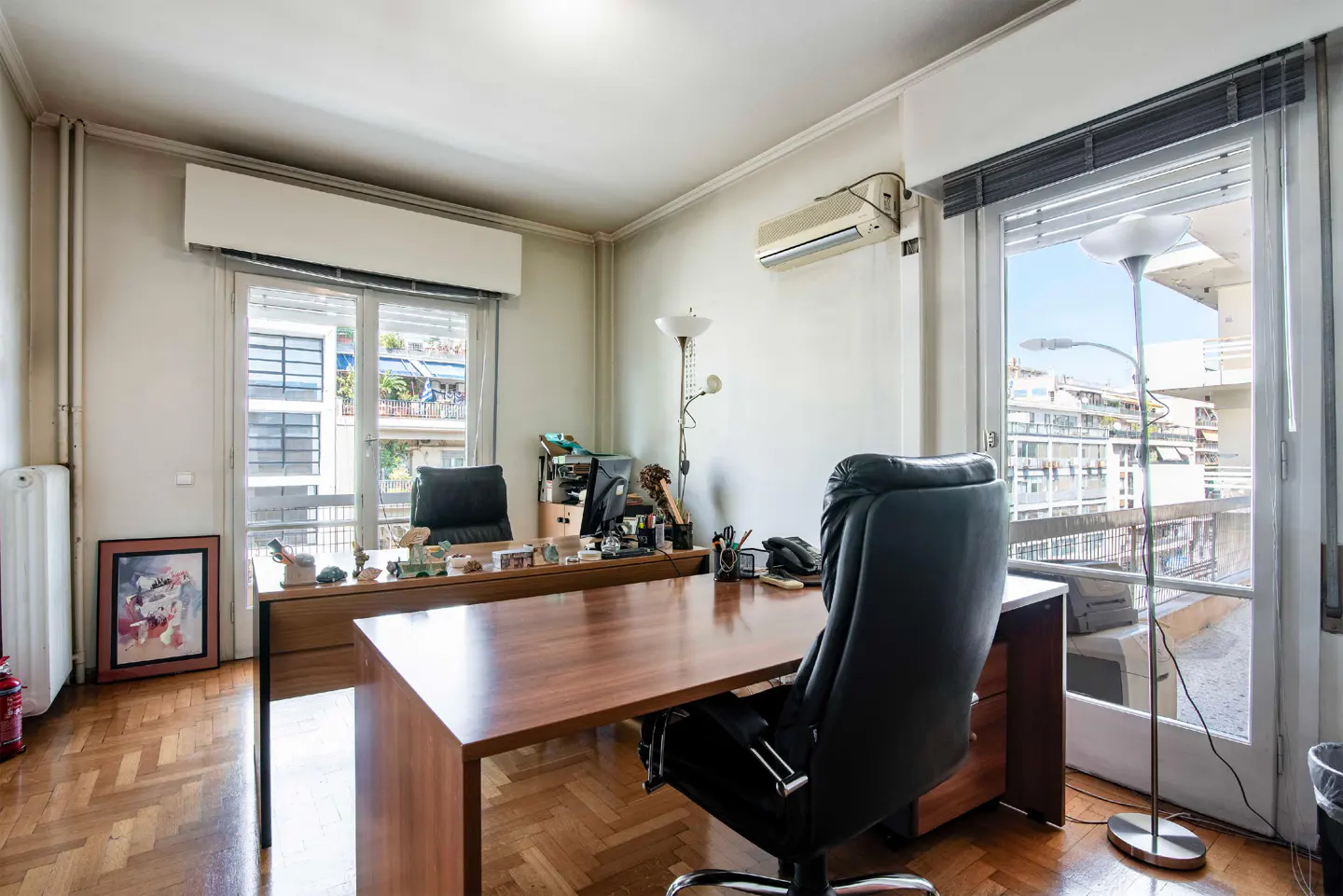 A bright office space with a large wooden desk, black leather chair, and windows overlooking a city view.