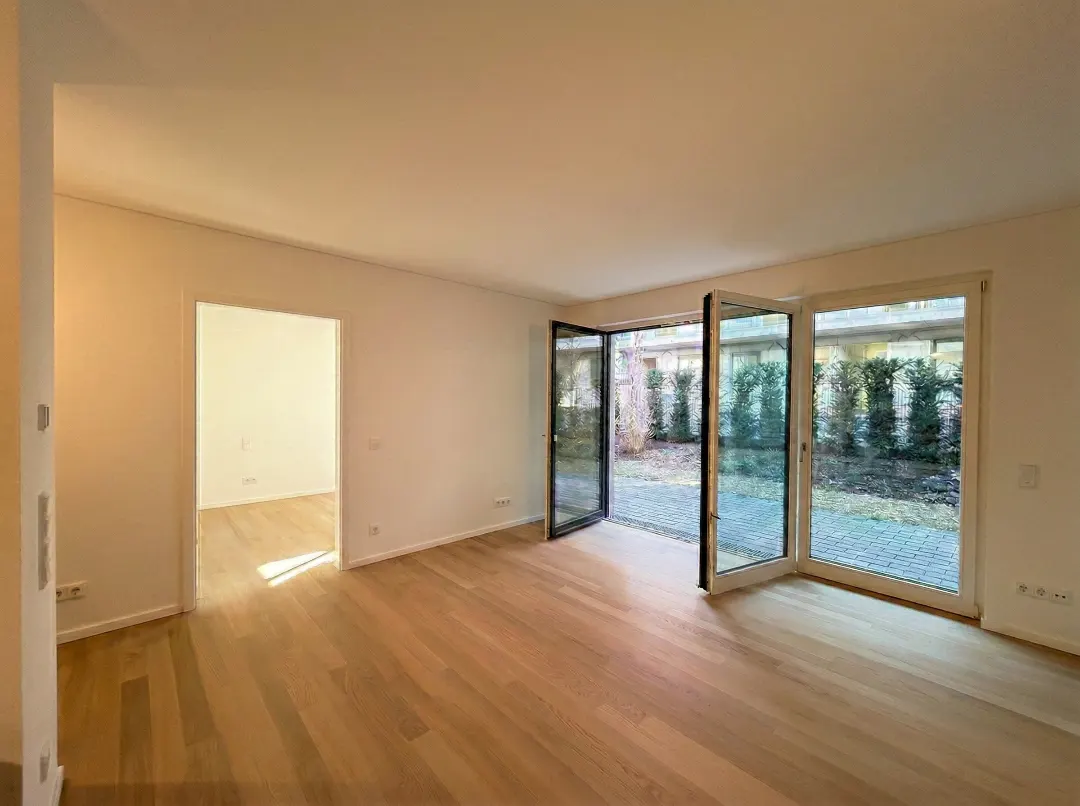 Bright, empty room with light wood floors and white walls. Glass doors open to a patio with green bushes. Doorway to another room on the left.