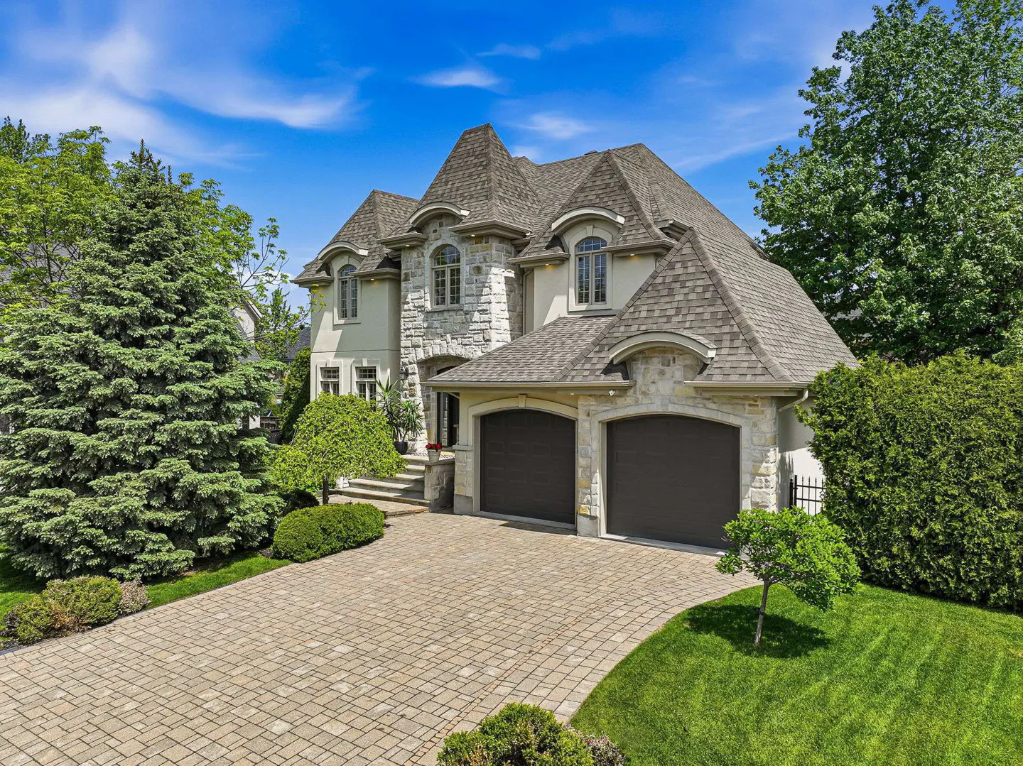 Two-story stone house with a double garage, paved driveway, and lush green landscaping under a bright blue sky.