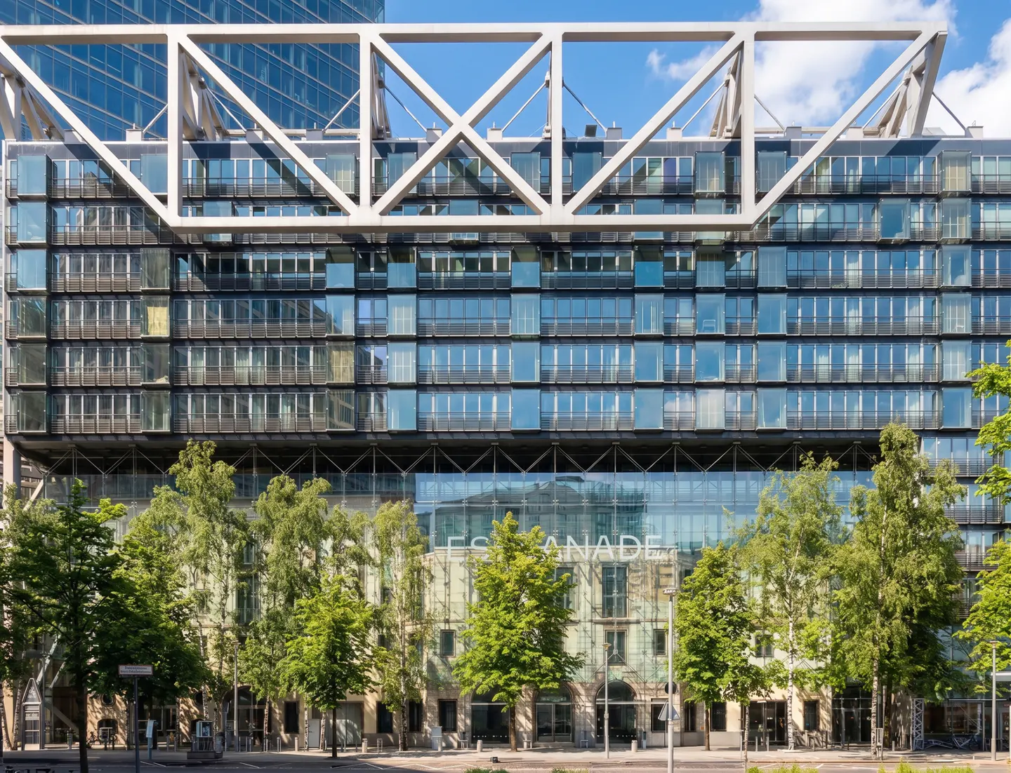 Modern office building with a glass facade, steel roof structure, and green trees in front under a blue sky.