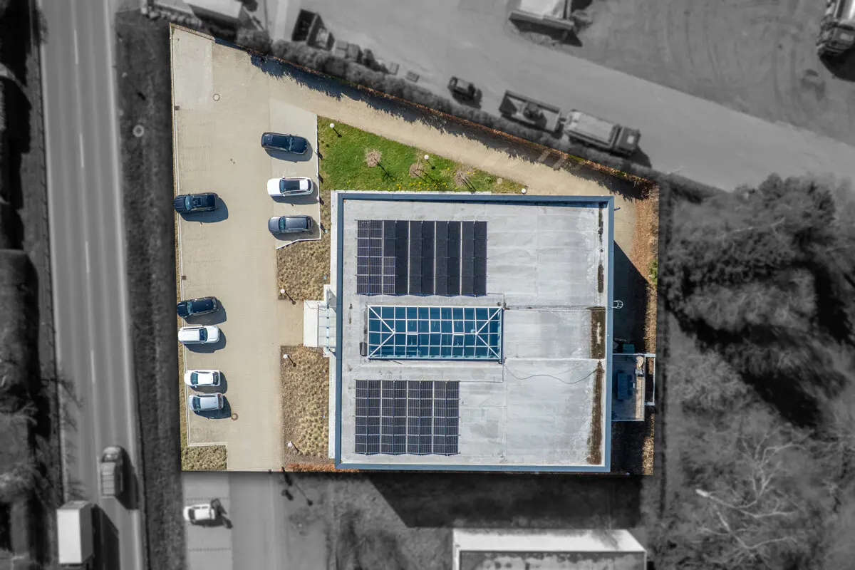 Aerial view of a building with solar panels and a skylight on its roof, next to a parking lot with cars.