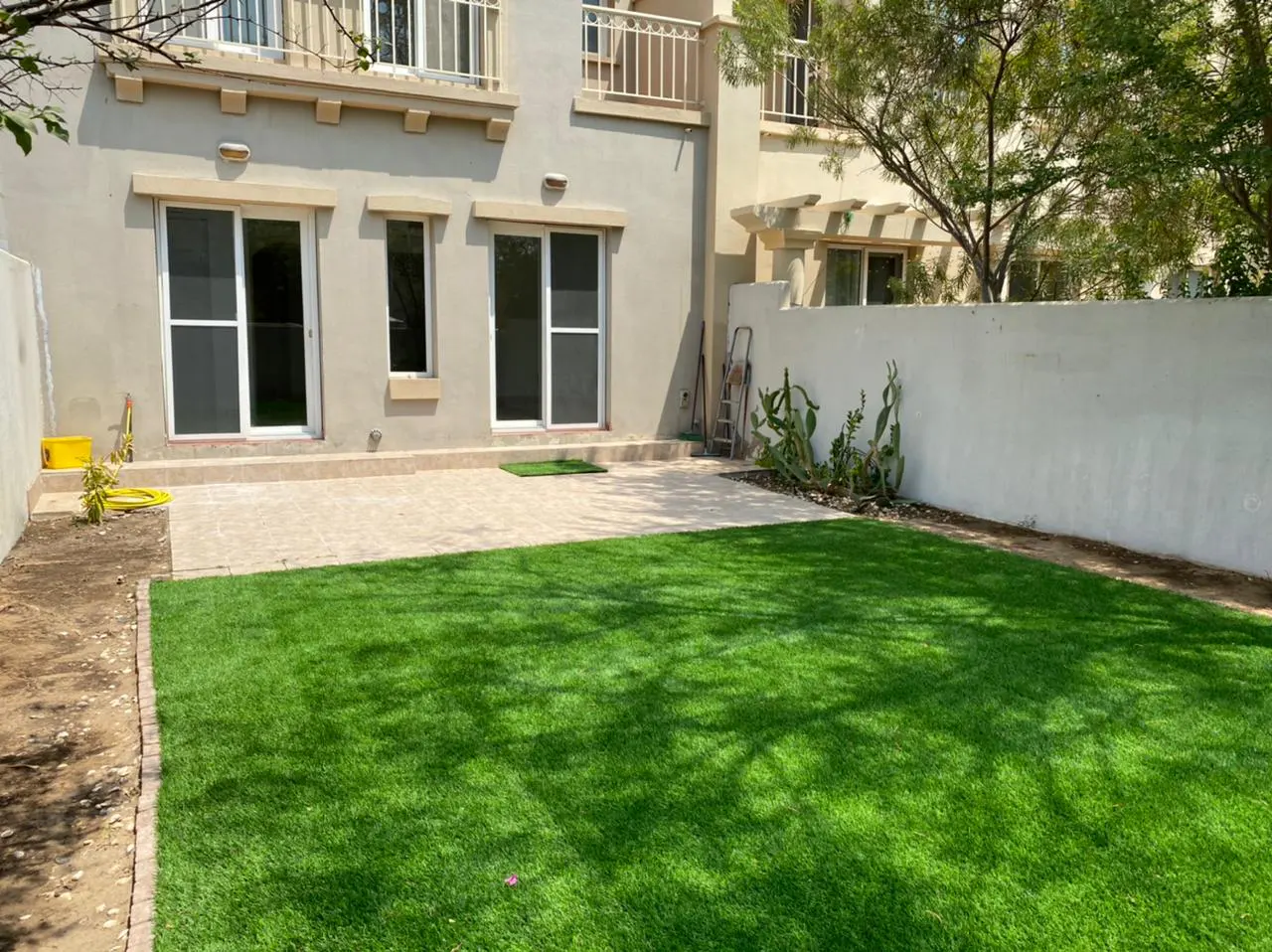 A backyard with green grass, a beige house with sliding glass doors, and a white wall with cacti.