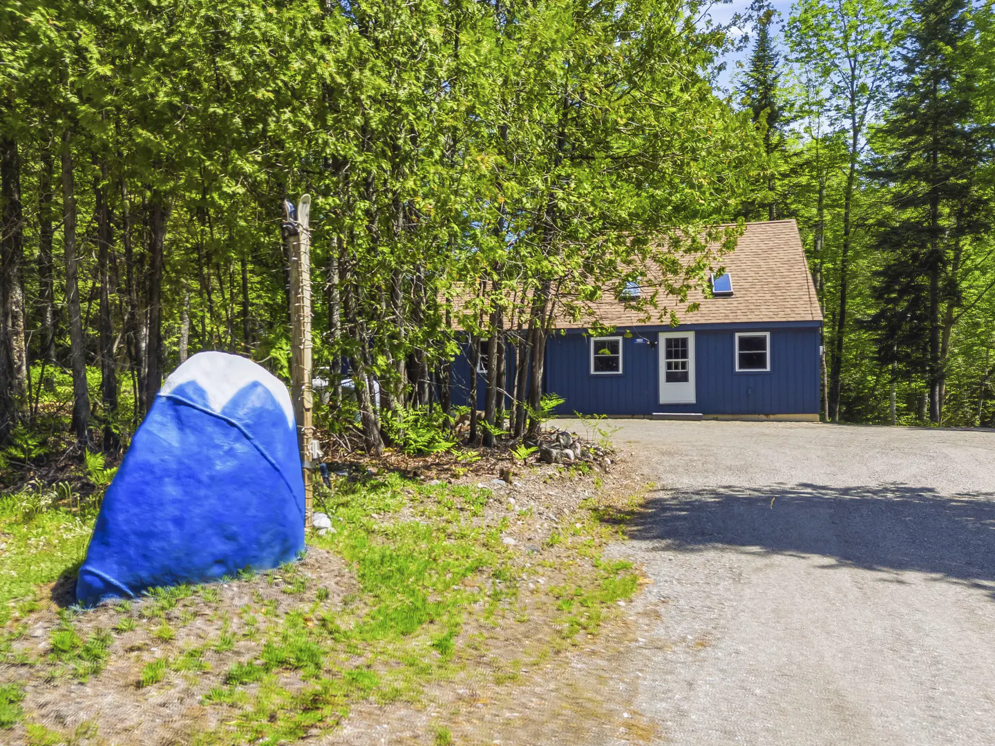 A blue cottage with a brown roof sits at the end of a gravel driveway, surrounded by lush green trees and a painted rock.