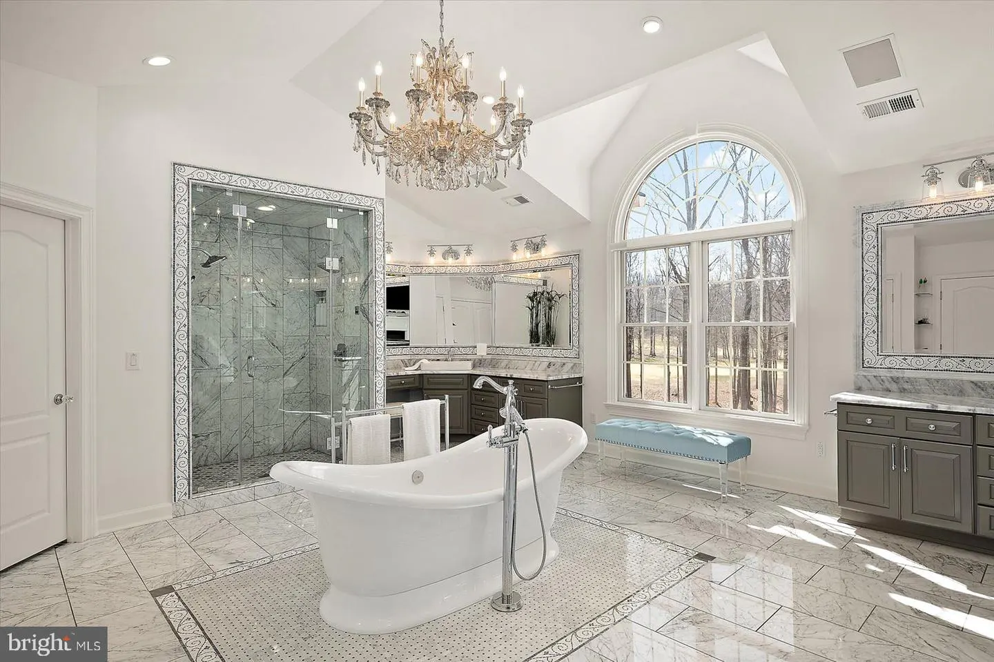 A luxurious bathroom with a white tub, marble floors, a glass shower, and a large arched window. A crystal chandelier hangs from the ceiling.