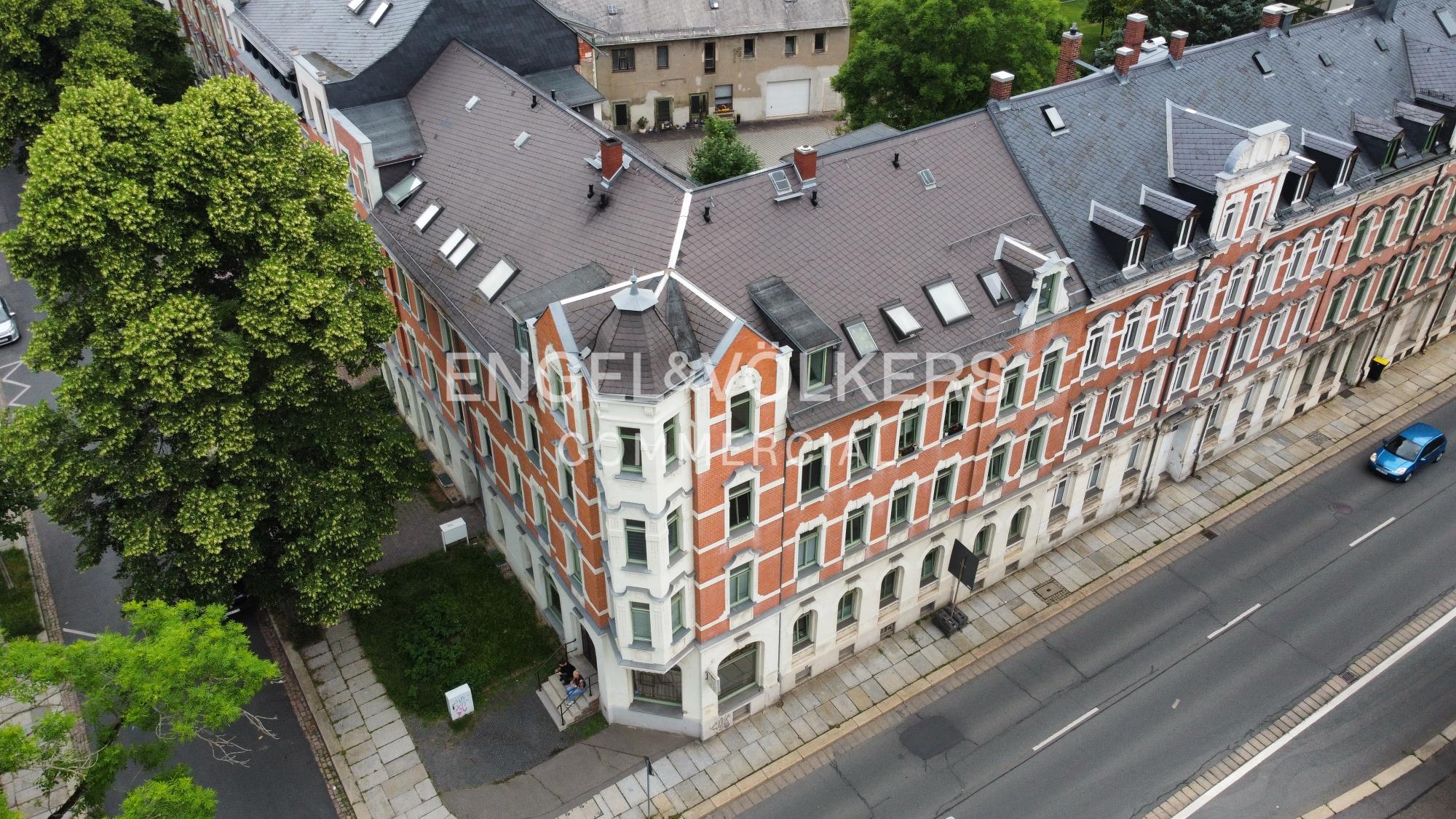 Aerial view of a multi-story red brick corner building with a dark roof, adjacent to a road and large green trees.