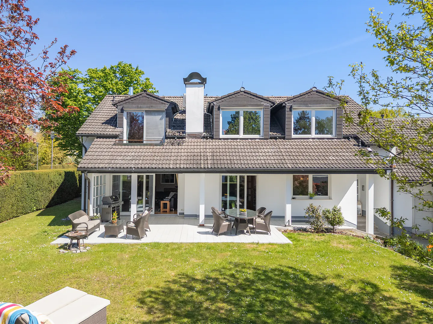 Rear view of a white house with a brown tile roof, dormer windows, and a patio with outdoor furniture on a green lawn.
