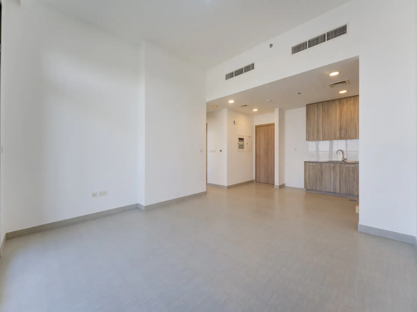 Empty apartment with white walls, gray floors, and a kitchen area with wood cabinets.