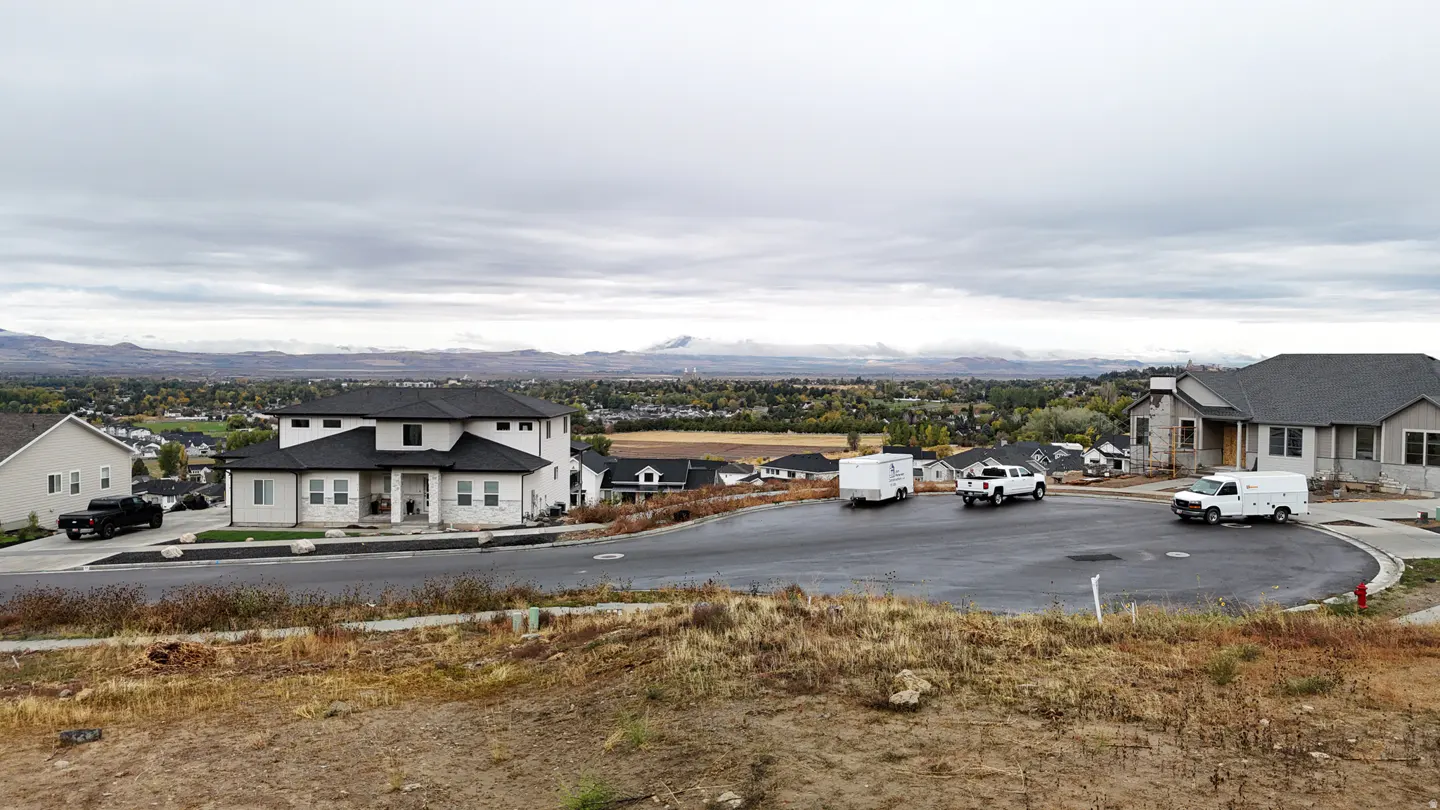 View of new homes on a cul-de-sac, with trucks and a trailer parked on the street. Mountains and a cloudy sky are in the background.