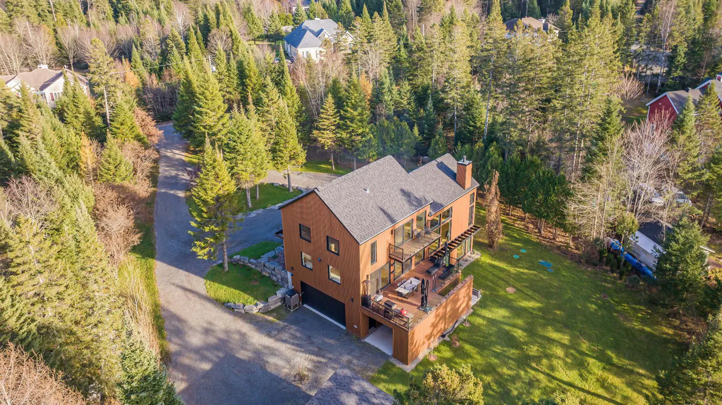 Aerial view of a modern brown house with a gray roof, surrounded by green trees and a lawn.