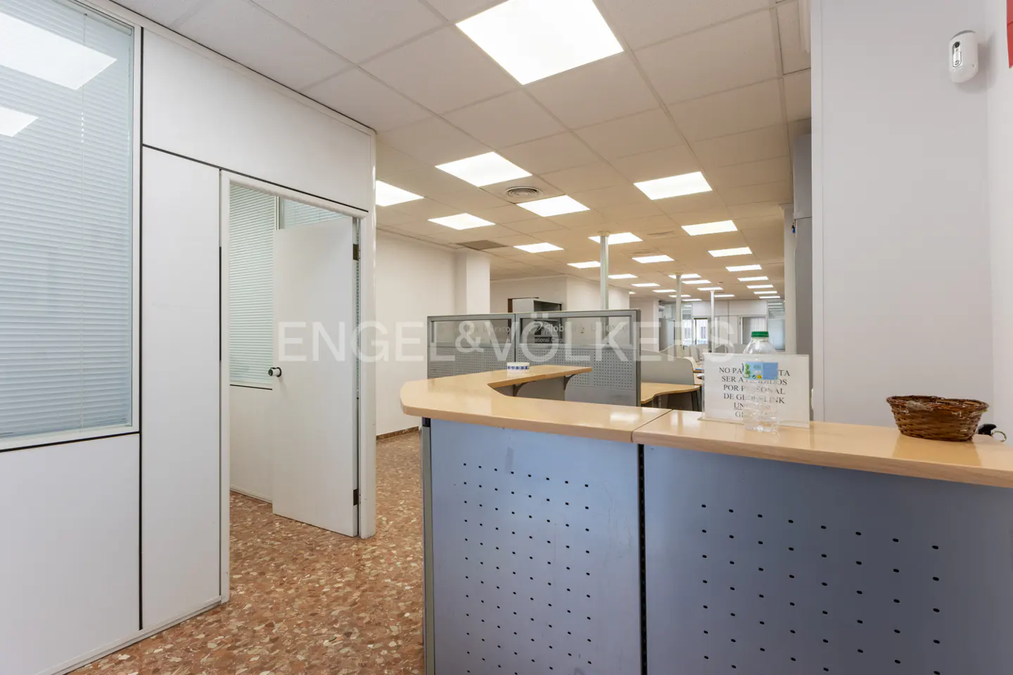 Office space with a reception desk, cubicles, and a door leading to another room. The floor is tiled in a speckled pattern.