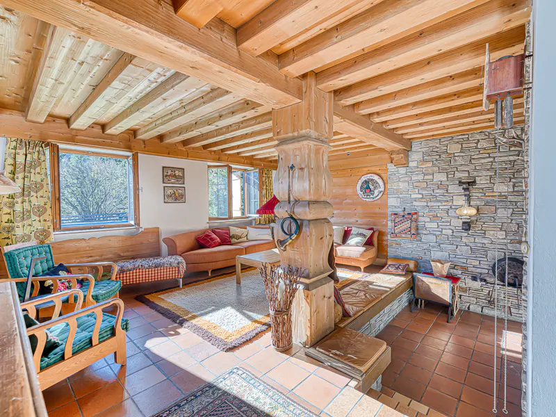 Living room with wood-beamed ceiling, stone wall, and terracotta tile floor. Sofas and chairs are arranged around a rug. Windows let in natural light.