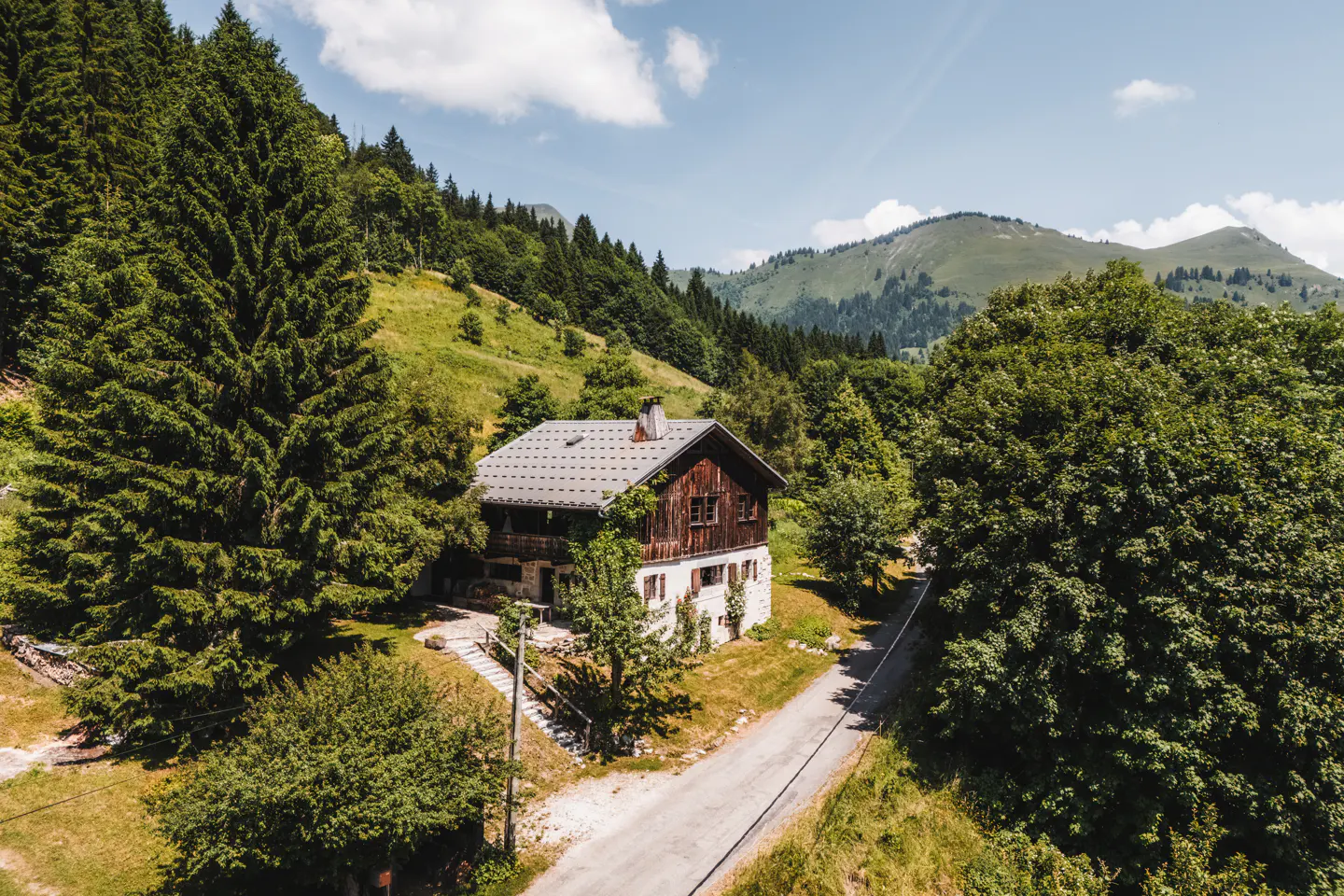 A two-story chalet with a gray roof sits nestled among green trees and rolling hills under a blue sky.