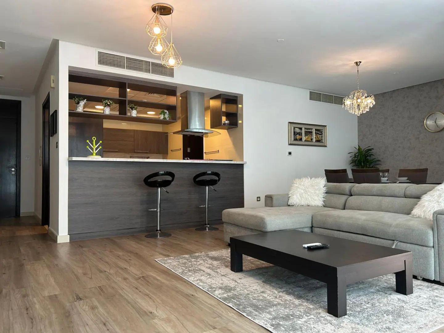 Open-concept living space with wood floors, a gray sectional sofa, and a black coffee table on a patterned rug. Kitchen with bar stools visible.