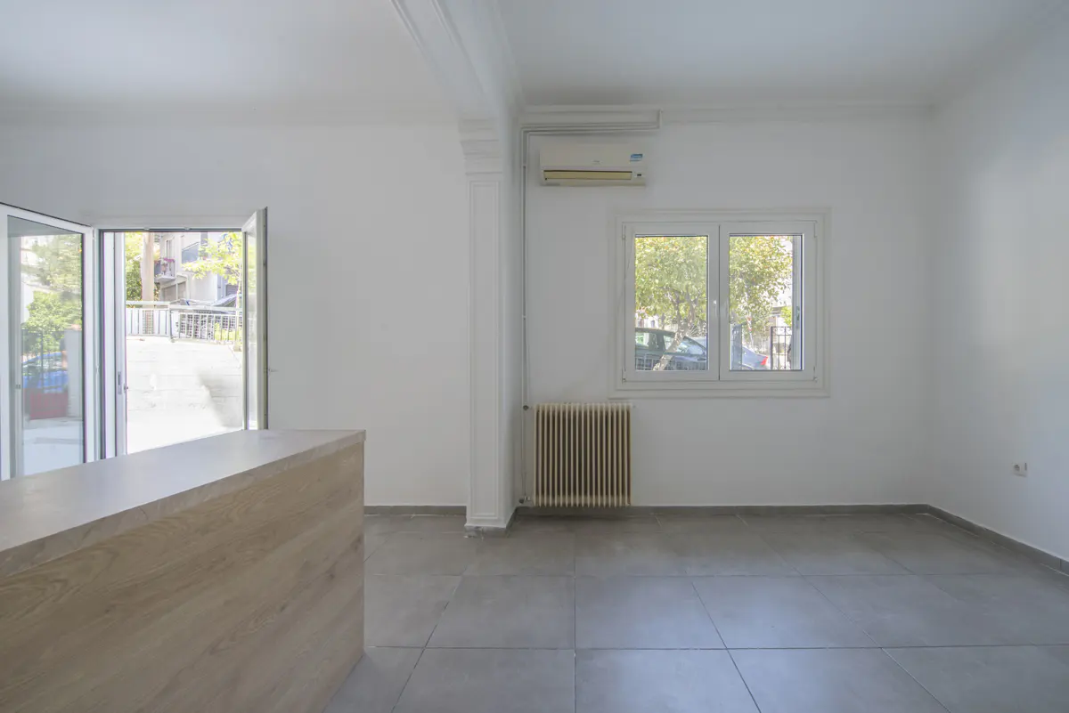 Bright, empty room with gray tile floor, white walls, and open glass doors to the outside. A wood-paneled counter is on the left.