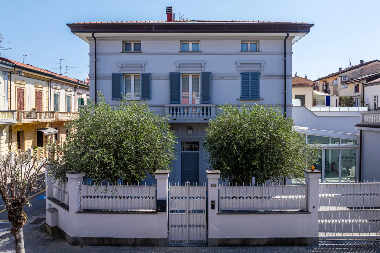 Two-story light blue house with blue shutters and a white fence and gate. Two green trees flank the gate.