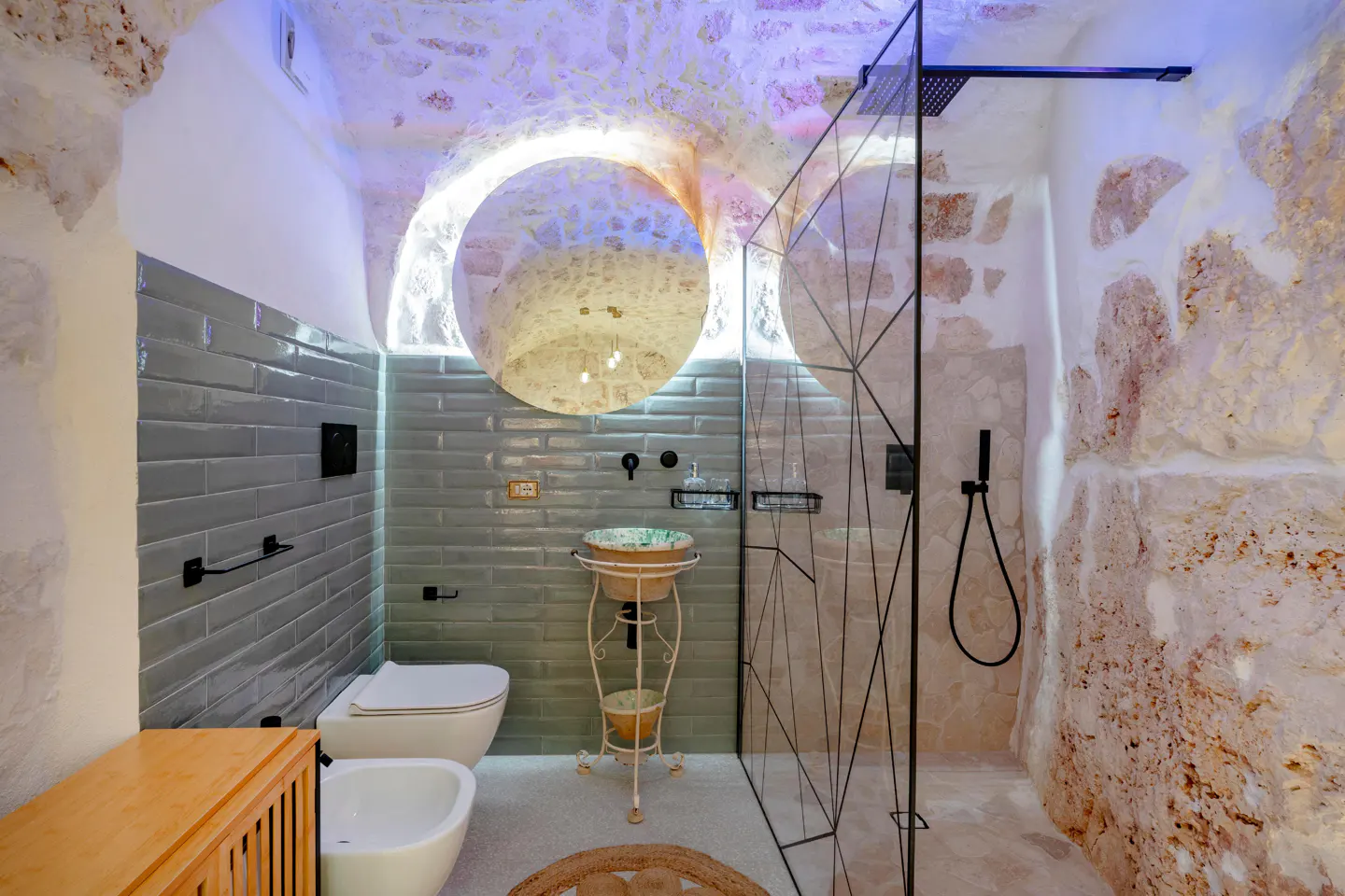 Bathroom with stone walls, blue-lit arched ceiling, gray tiled wall, toilet, bidet, sink on stand, and glass shower with black fixtures.