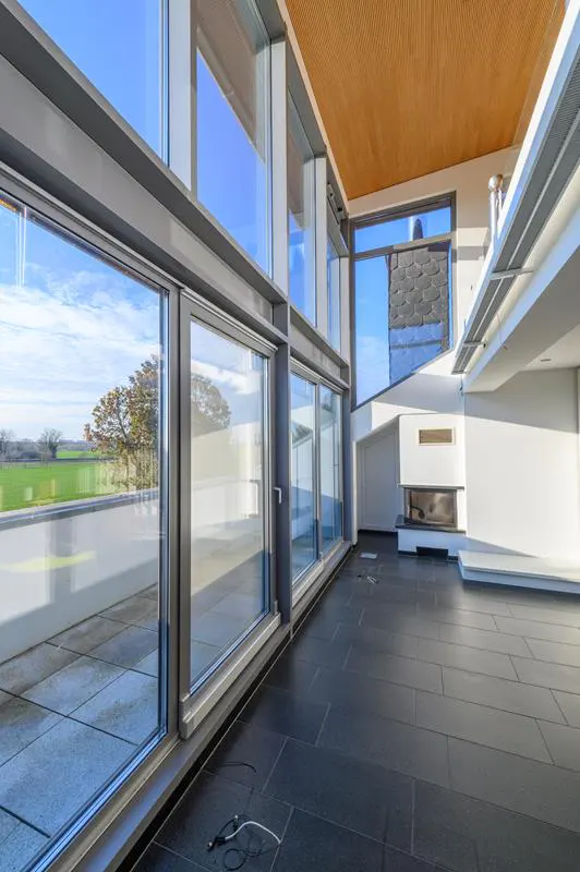 Bright, modern living room with black tile floor, wood ceiling, and large windows overlooking a green field. A fireplace is visible.