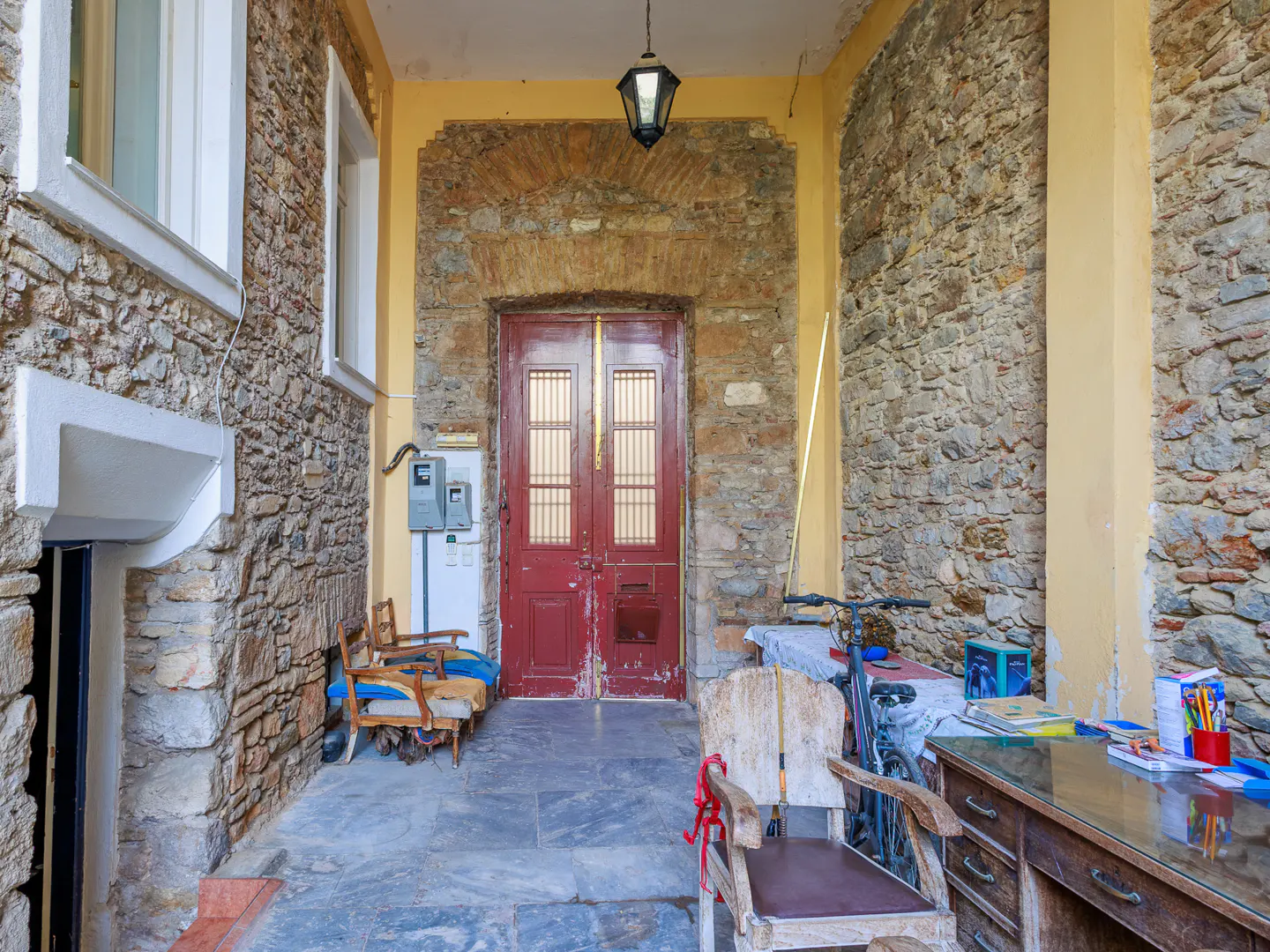Stone porch with a red door, stone walls, and yellow trim. A desk, chairs, and a bicycle are on the porch.