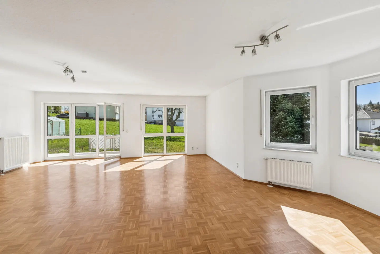 Bright, empty room with parquet floors, white walls, and large windows overlooking a green lawn and houses.