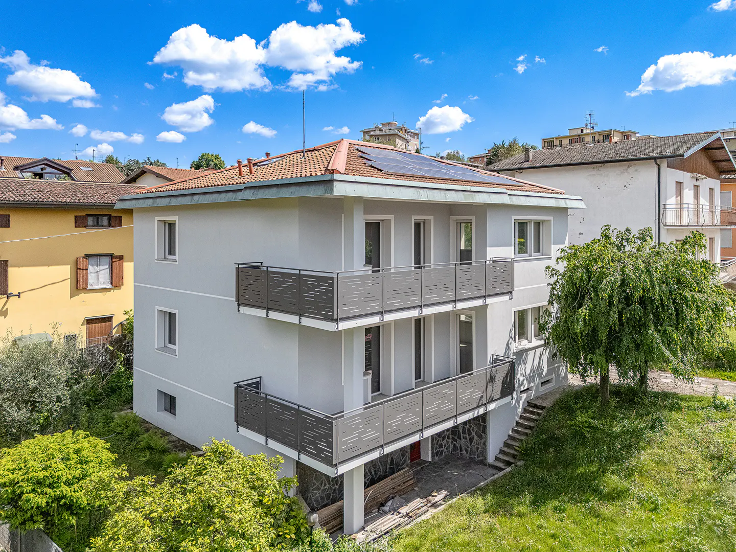 Exterior view of a three-story gray house with two balconies, a red tile roof, and solar panels under a blue sky.