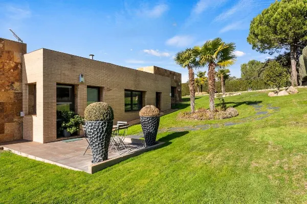 Modern tan brick home with a patio, green lawn, palm trees, and blue sky. Two large black planters sit on the patio.