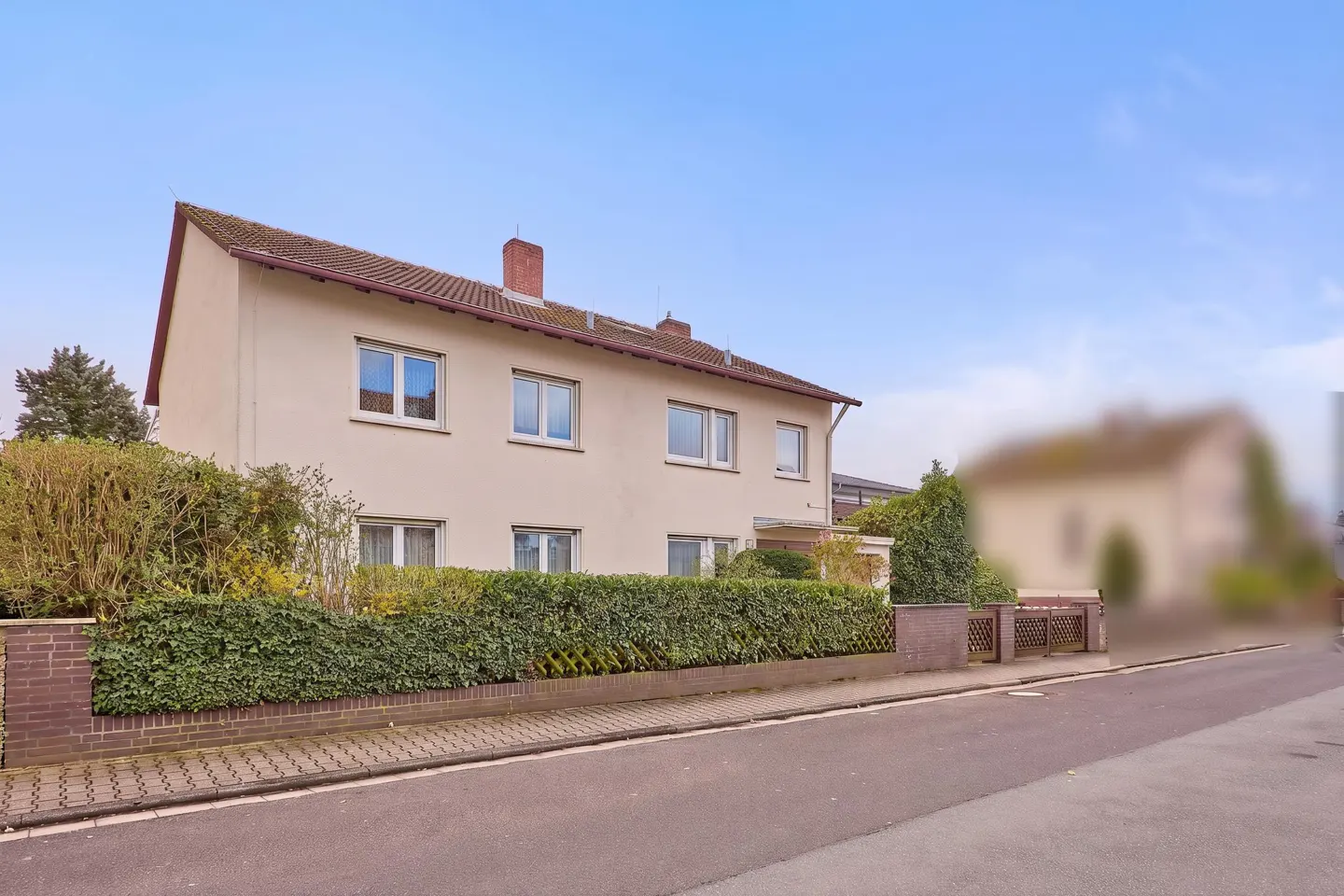 Two-story beige house with a brown roof and chimney, green hedge, and brick fence under a blue sky.