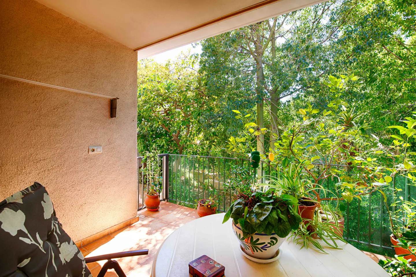 A sunny balcony with a white table, potted plants, and a chair with a floral cushion overlooks lush green trees.