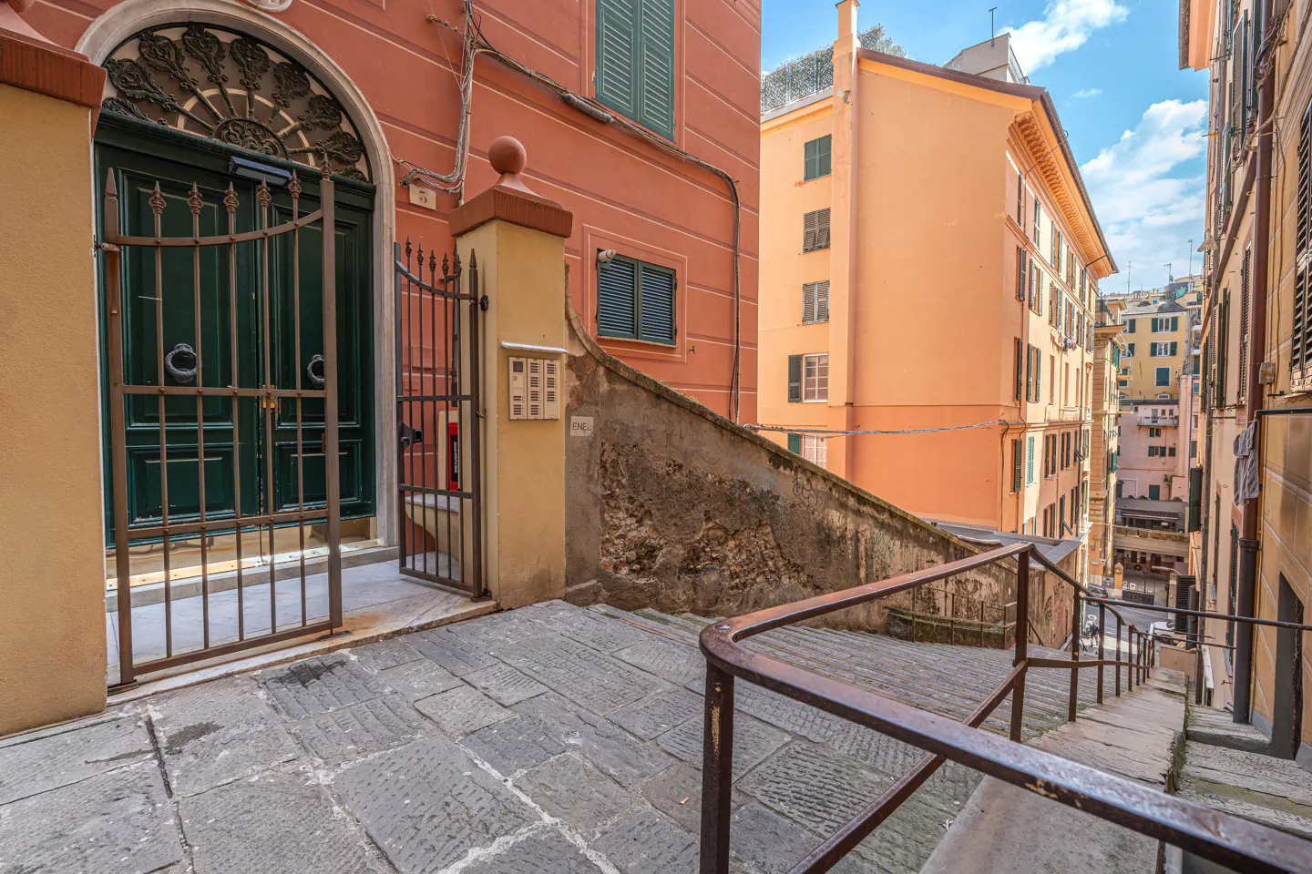 Exterior view of a building with a green door and metal gate, stone steps, and orange buildings in the background.