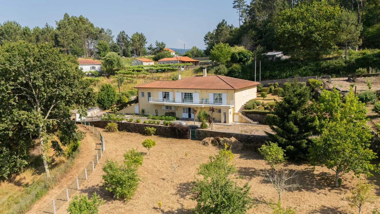 A two-story yellow house with a red tile roof, white trim, and a balcony, surrounded by trees and a brown field.